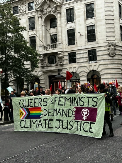 a group of people walking down a street holding a banner, by Benjamin Block, pexels contest winner, feminist art, wet climate, london streets in background, lesbian, background image