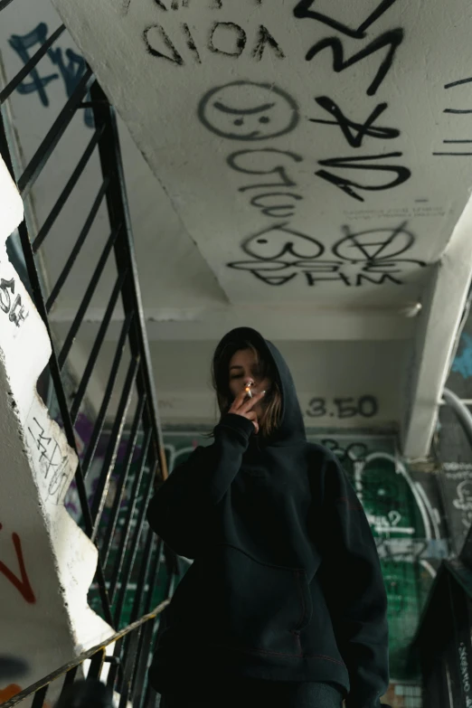 a woman in a black hoodie standing on an escalator, inspired by Elsa Bleda, trending on pexels, graffiti, outside alone smoking weed, ( ( theatrical ) ), goth girl aesthetic, on a wall