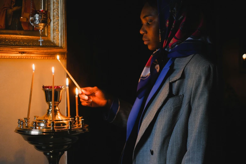 a woman holding a lit candle in a dark room, standing in front of the altar, somali woman, profile image, wearing a grey robe