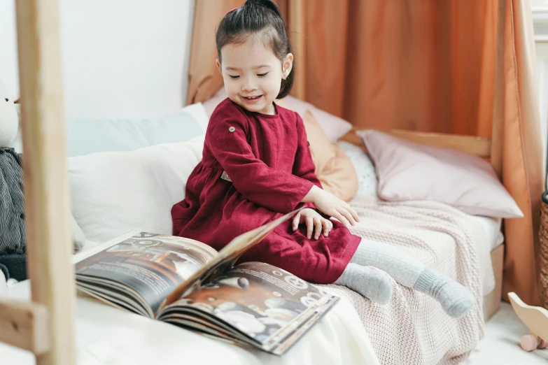 a little girl sitting on a bed reading a book, pexels contest winner, crimson clothes, korean girl, science magazines, organic dress