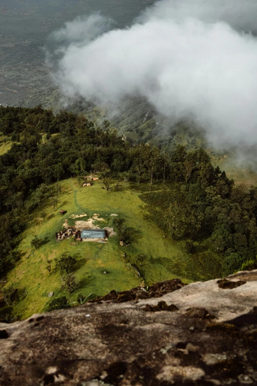 a man standing on top of a rock next to a lush green hillside, a tilt shift photo, happening, tribe huts in the jungle, view above the clouds, grey, helipad