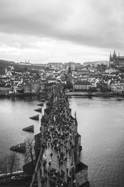 a black and white photo of people walking across a bridge, a black and white photo, renaissance, prague in the background, drone view of a city, full of people, bridge over the water