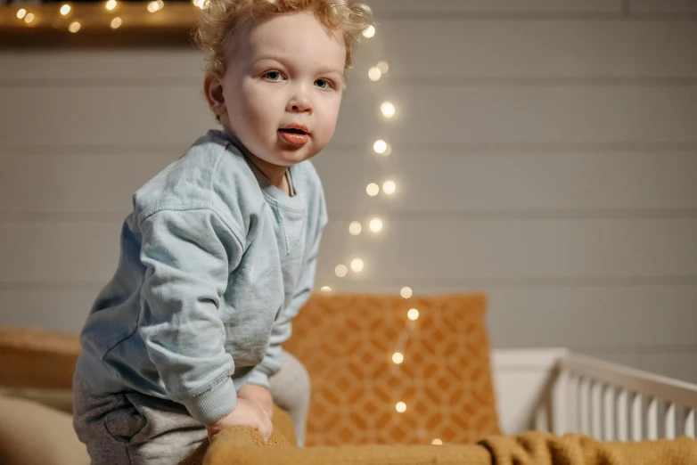 a baby sitting in a crib with christmas lights in the background, a portrait, by Jakob Gauermann, pexels contest winner, visual art, blond boy, sitting on couch, light cute freckles, doing a sassy pose