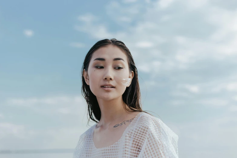 a woman standing on top of a beach next to the ocean, inspired by Ai Xuan, trending on pexels, hyperrealism, beauty mark on cheek, ethnicity : japanese, wearing translucent sheet, cumulus tattoos