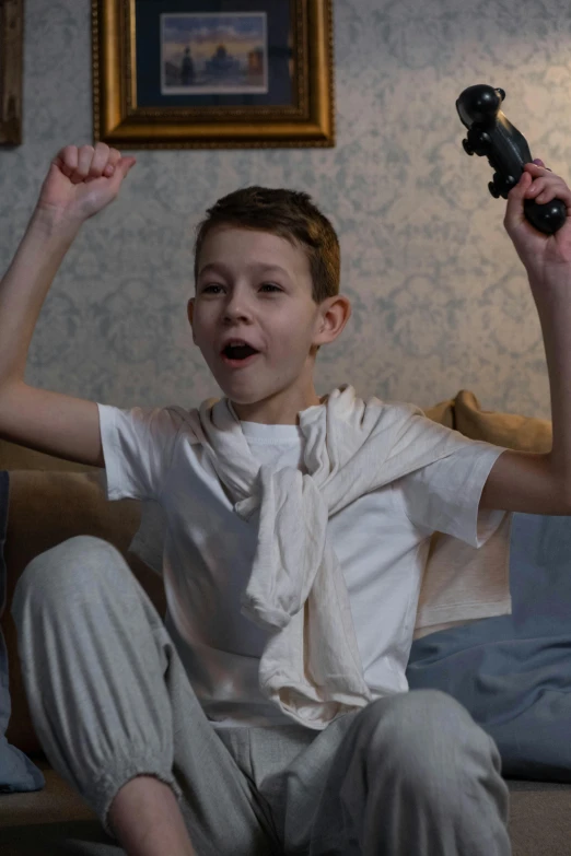 a young boy sitting on a couch holding a remote control, inspired by Samuel Colman, happening, fists in the air, with a gun, wearing white pajamas, video still