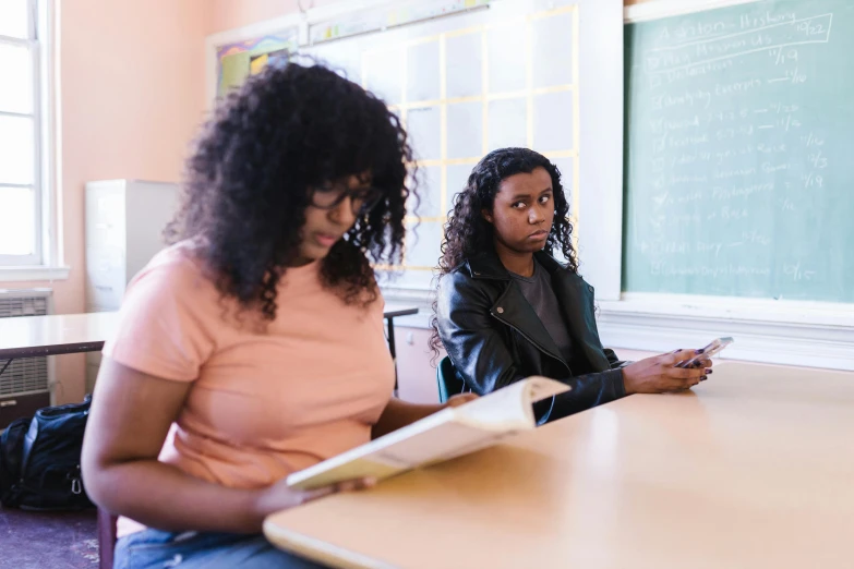 two women sitting at a table in front of a chalkboard, by Carey Morris, trending on unsplash, figuration libre, standing in class, black teenage girl, 15081959 21121991 01012000 4k, trying to read