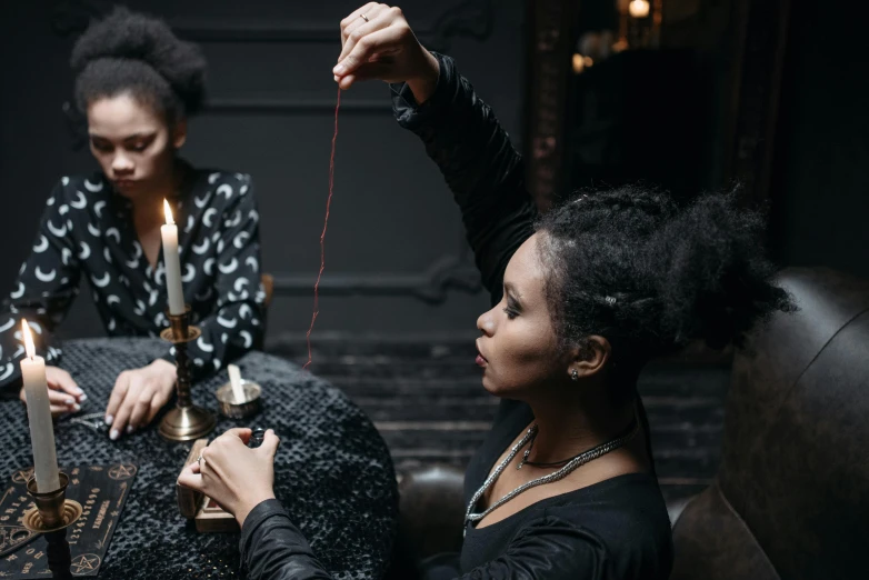 two women sitting at a table with candles, pexels contest winner, black arts movement, holding a magic needle, black and red silk clothing, holding electricity, haunted