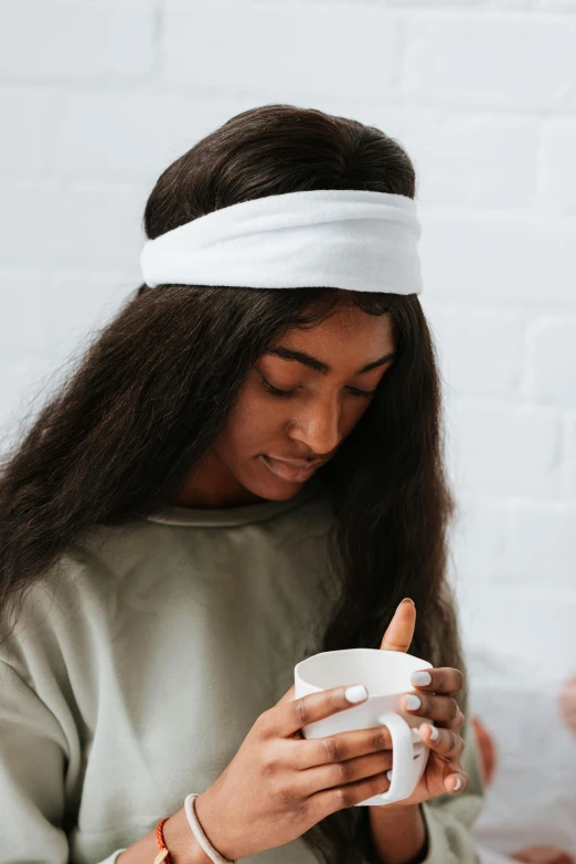 a woman sitting on a bed holding a cup of coffee, wearing the number 1 headband, white hue, loosely cropped, head and shoulders view