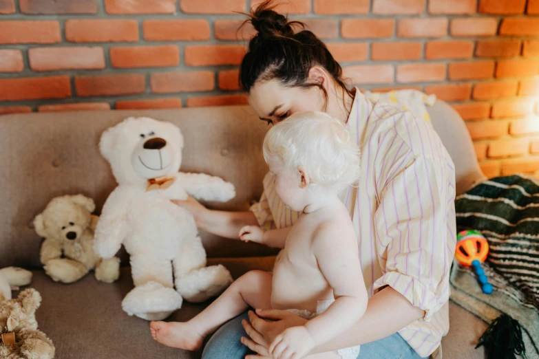 a woman sitting on a couch holding a baby next to a teddy bear, pexels contest winner, white, kids toys, profile image, 1 2 9 7