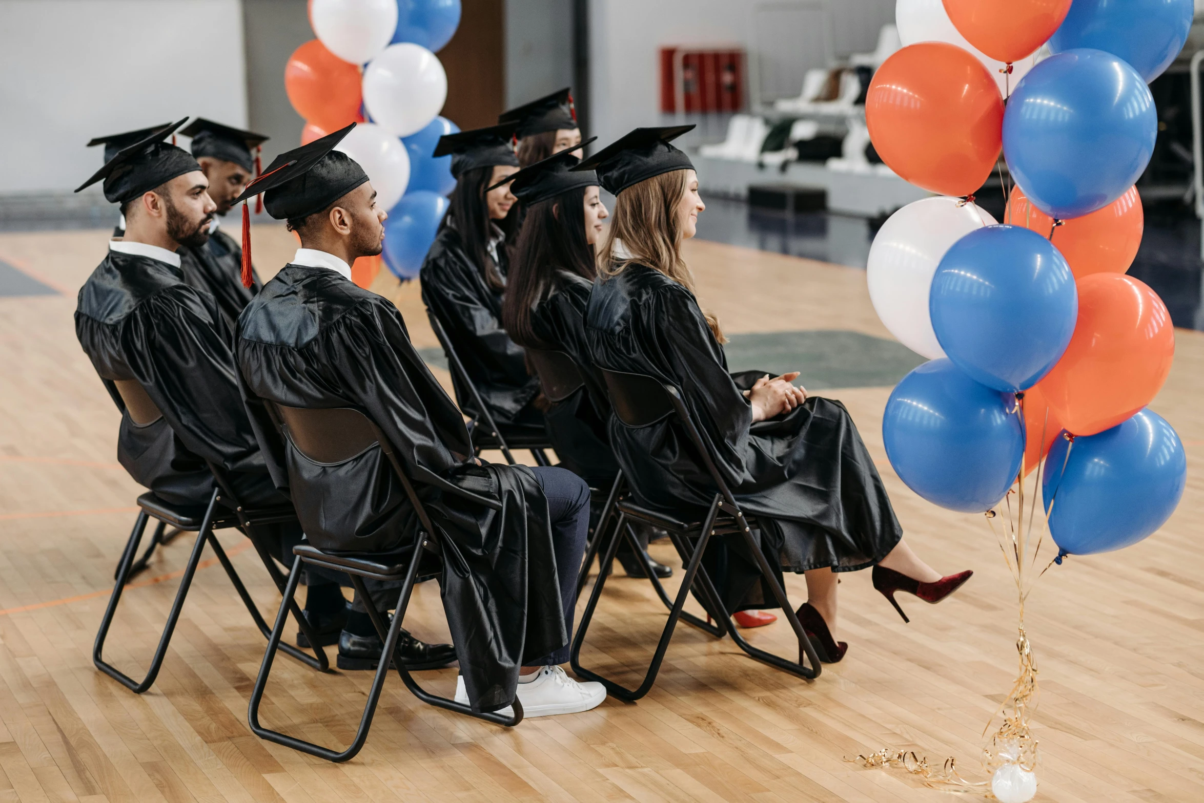 a group of people sitting on top of a wooden floor, wearing an academic gown, baloons, trending on attestation, sitting on a chair