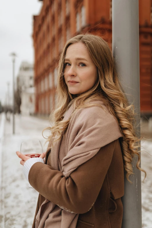 a woman leaning against a pole in the snow, inspired by Louisa Matthíasdóttir, pexels contest winner, renaissance, light brown long wavy hair, brown jacket, gif, modest