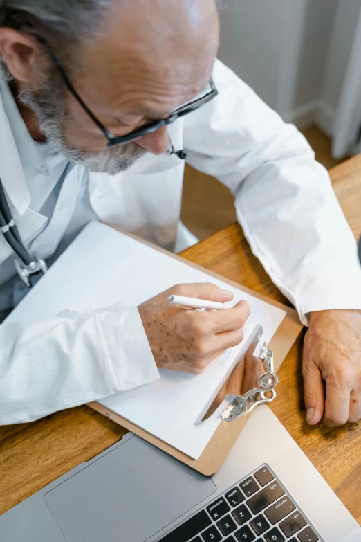 a man sitting at a desk in front of a laptop computer, by Adam Marczyński, pexels contest winner, wearing a white hospital gown, writing on a clipboard, white reading glasses, a high angle shot