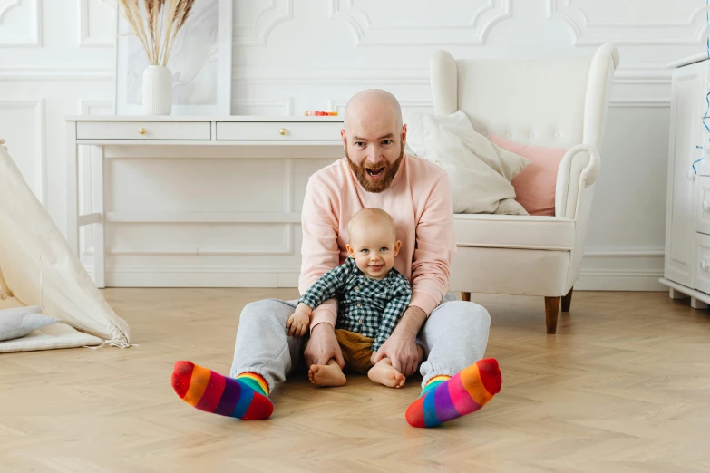 a man sitting on the floor holding a baby, pexels contest winner, rainbow clothes, bald, bearded, striped socks