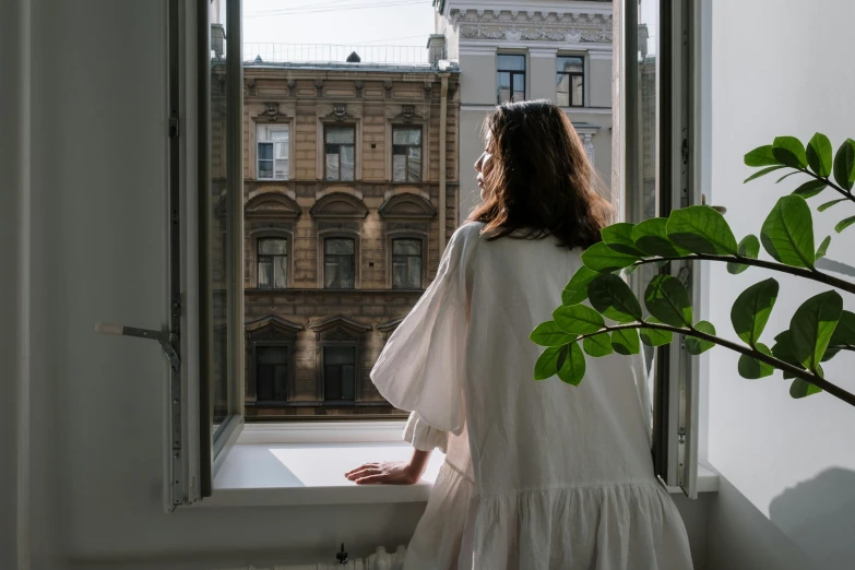 a woman in a white dress looking out a window, pexels contest winner, city apartment cozy calm, neo kyiv, wearing a baggy pajamas, from back