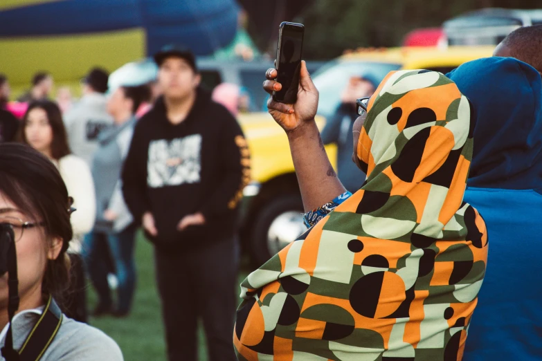 a woman taking a picture of a crowd of people, unsplash, wearing an african dress, hood and cape, at a park, te pae