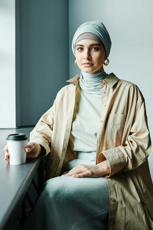 a woman sitting at a table with a cup of coffee, inspired by Maryam Hashemi, hurufiyya, wearing casual clothing, promo image, multilayered outfit, looking serious