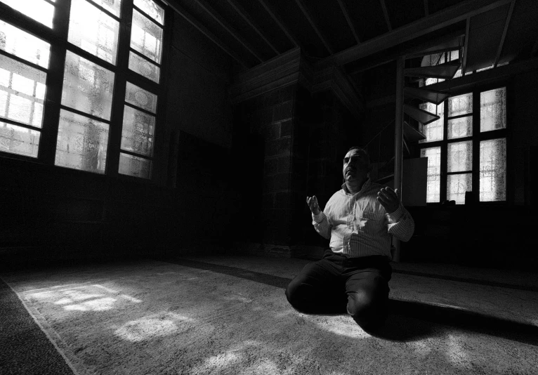 a black and white photo of a man sitting on the floor, inspired by Abdullah Gërguri, hurufiyya, séance, kneeling at the shiny floor, harsh sunlight, uploaded