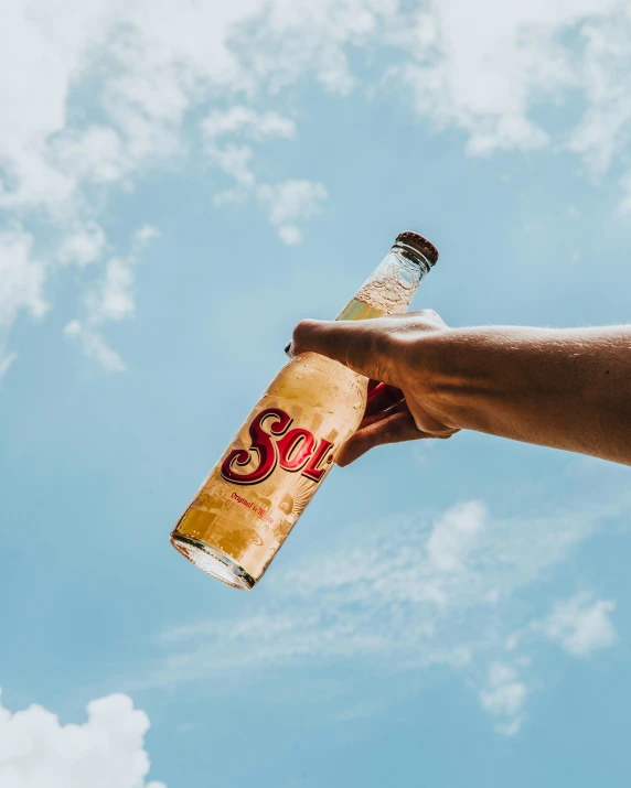 a hand holding a bottle of soda against a blue sky, by Samuel Scott, pexels contest winner, sots art, light tan, 🍸🍋, sol retreat, view from ground