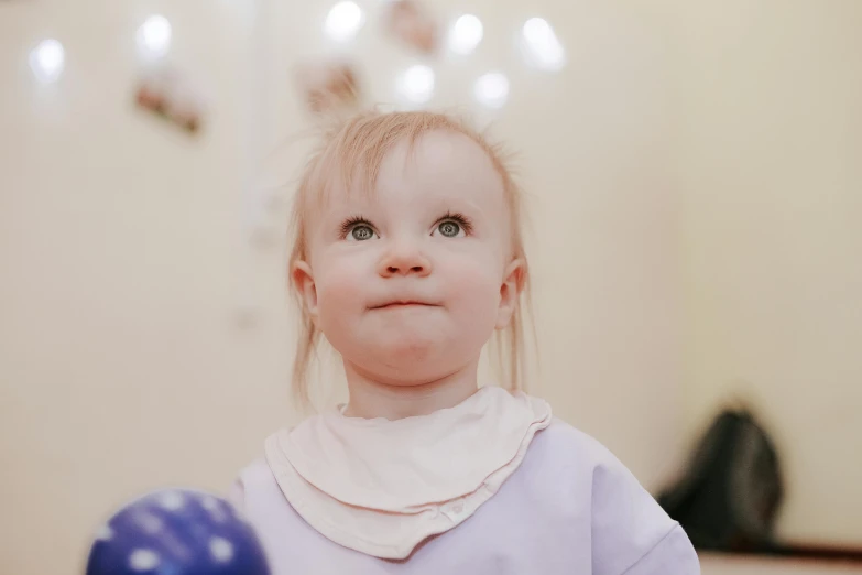 a little girl holding a blue ball in front of her face, by Emma Andijewska, pexels contest winner, as she looks up at the ceiling, pale skin and purple eyes, soft light.4k, cleft chin