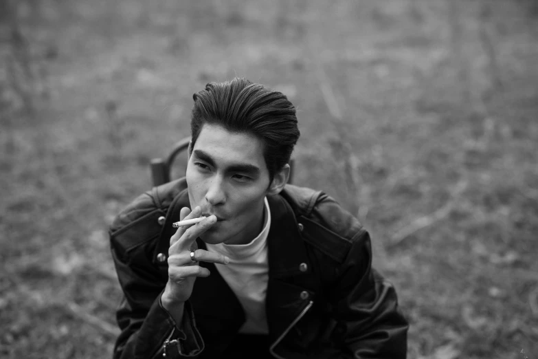 a black and white photo of a man smoking a cigarette, by Emma Andijewska, cai xukun, in an old 1950s leather jacket, sitting in a field, modeling shoot