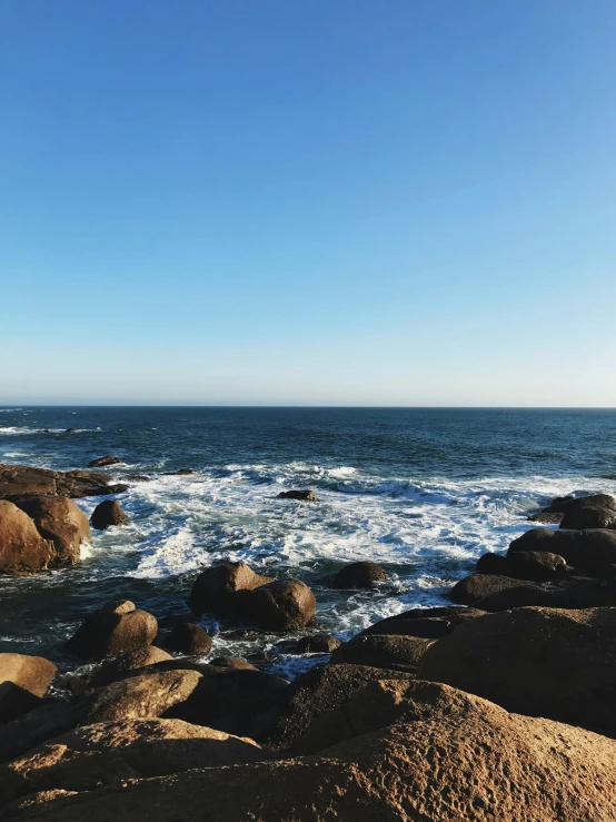 a man standing on top of a rock next to the ocean, clear skies in the distance, boulders, ocean spray, profile image