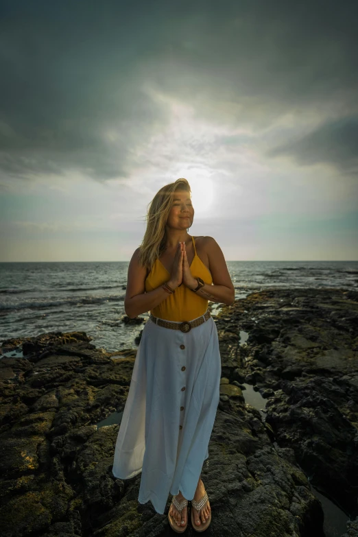 a woman standing on top of a rock next to the ocean, an album cover, unsplash, renaissance, lotus pose, sun lighting, blonde goddess, yellow