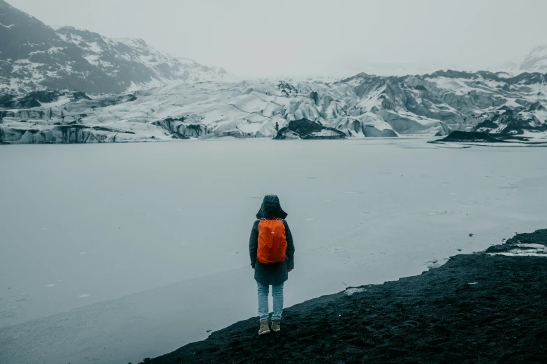 a person standing on a rock near a body of water, by Louisa Matthíasdóttir, pexels contest winner, with a backpack, with a snowy mountain and ice, black sand, dark grey and orange colours