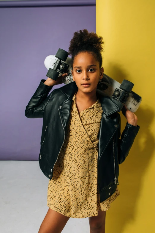 a young girl with a skateboard leaning against a yellow wall, trending on unsplash, wearing leather jacket, looking to camera, kids, studio lighting”