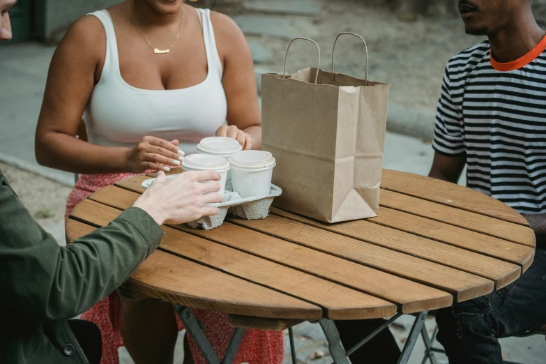 a group of people sitting around a wooden table, the handbag is over a wood table, paper cup, wearing a sexy cropped top, at the park