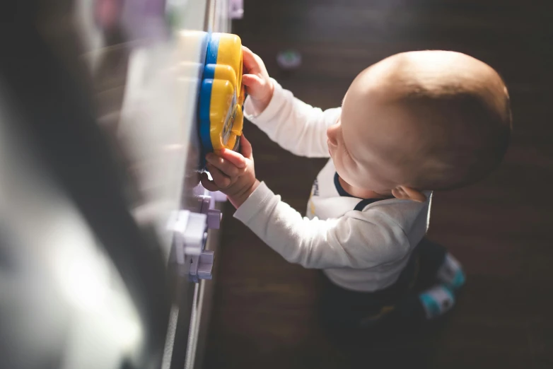 a baby playing with a toy in front of a refrigerator, by Matthias Stom, pexels contest winner, happening, tactile buttons and lights, holding a shield, high angle close up shot, standing on a shelf