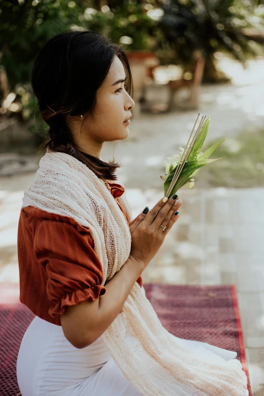 a woman sitting on top of a red and white rug, inspired by Ruth Jên, unsplash, renaissance, cambodia, blessing palms, holding a wood piece, exterior