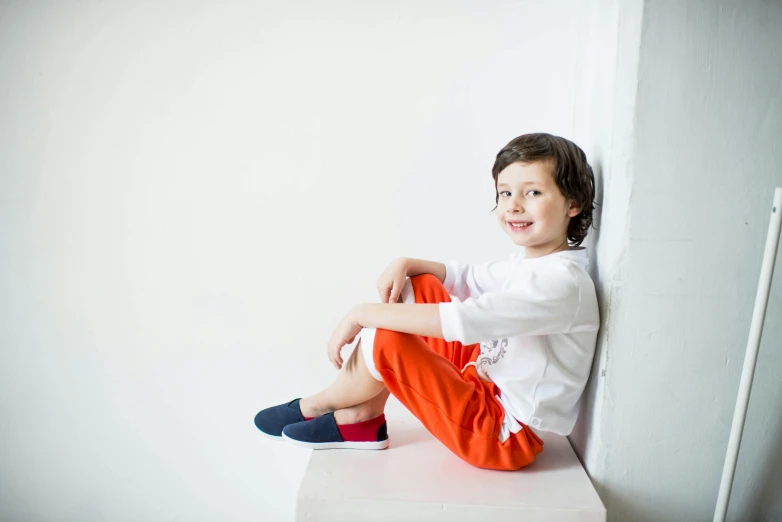 a little boy sitting on top of a white table, by Nina Hamnett, blue long pants and red shoes, white and orange, official store photo, leaning against the wall