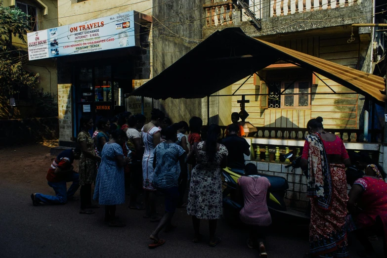 a group of people standing around a food cart, praying, stood outside a corner shop, distant photo