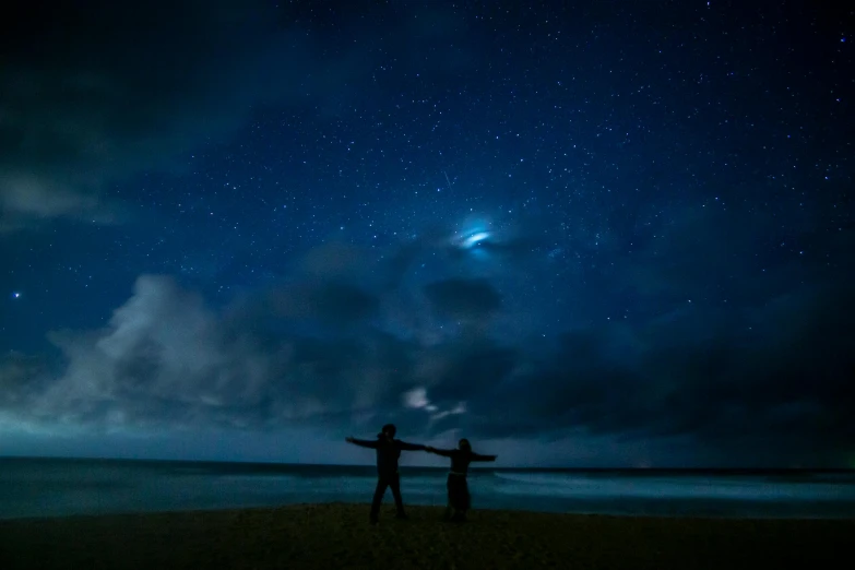 two people standing on a beach under a night sky, by Jessie Algie, pexels contest winner, hurufiyya, dramatic blue lighting, pointing to heaven, ( ( ( ( kauai ) ) ) ), search lights