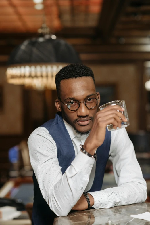 a man sitting at a bar drinking a glass of water, inspired by Barthélemy Menn, trending on unsplash, square rimmed glasses, ( ( dark skin ) ), confident pose, elegantly dressed