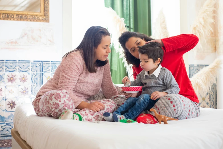 a woman and two children sitting on a bed, pexels contest winner, red sweater and gray pants, spanish, multicoloured, holiday