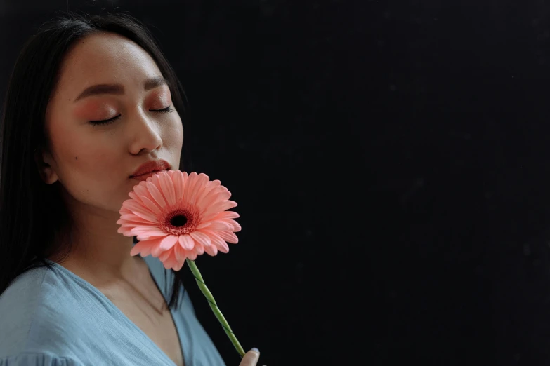 a woman holding a pink flower in front of her face, inspired by Itō Shinsui, pexels contest winner, standing with a black background, vietnamese woman, holding daisy, coral lipstick