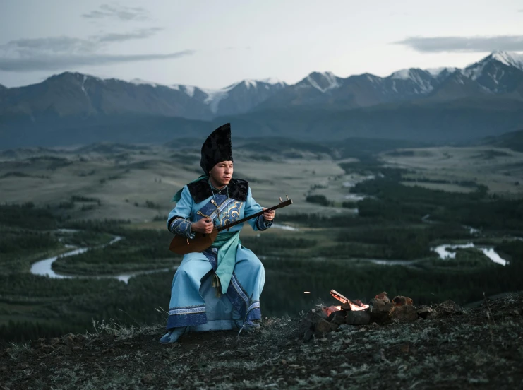 a man sitting on top of a hill next to a fire, an album cover, inspired by Konstantin Vasilyev, pexels contest winner, hurufiyya, traditional dress, instrument, national geographic photo award, singing