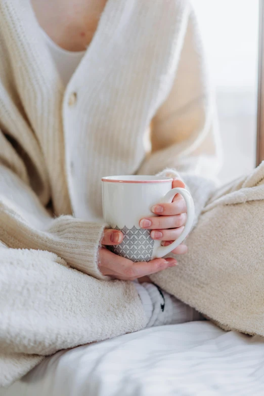 a woman sitting on a bed holding a cup of coffee, trending on pexels, sitting with wrists together, cold weather, beige, manuka