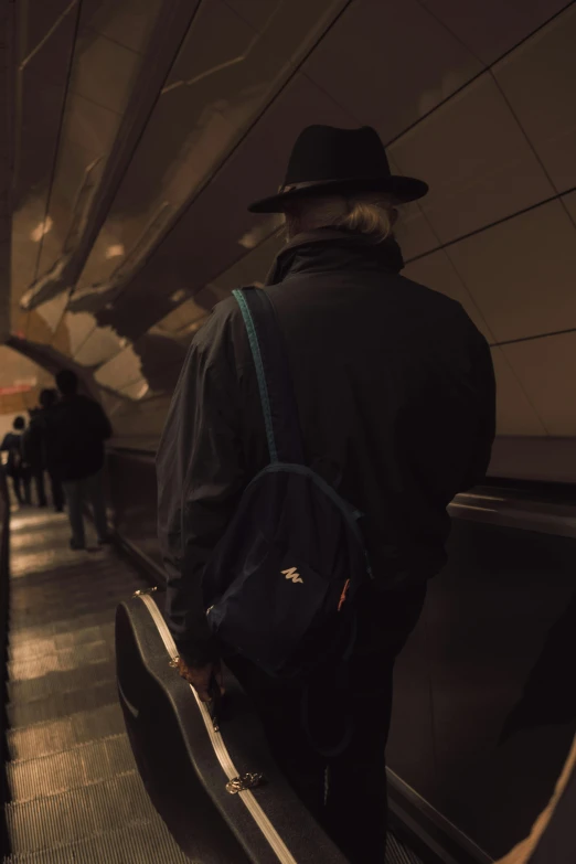 a man in a hat is riding an escalator, an album cover, unsplash, carrying a saddle bag, low light cinematic, pondering, alexey gurylev