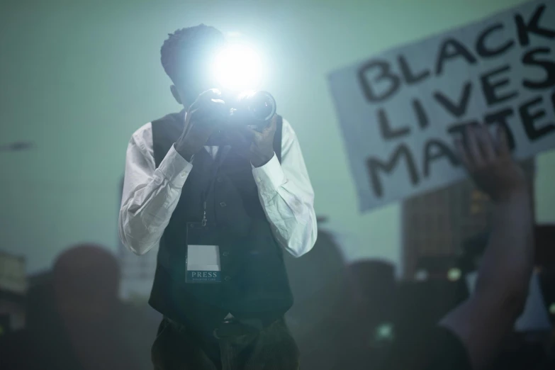 a man holding a camera in front of a crowd, black arts movement, bleached strong lights, ap news, concept photo, bl