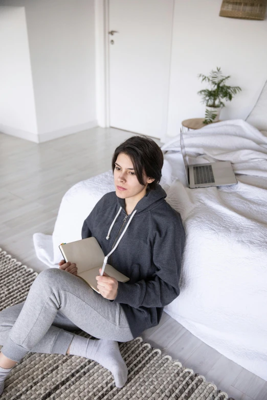 a woman sitting on a bed reading a book, grey hoodie, holding notebook, looking serious, 2019 trending photo