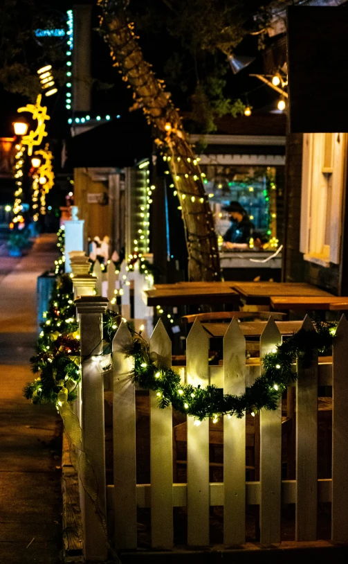 a street filled with lots of lights next to a wooden fence, in a sidewalk cafe, decorations, ivy's, holiday