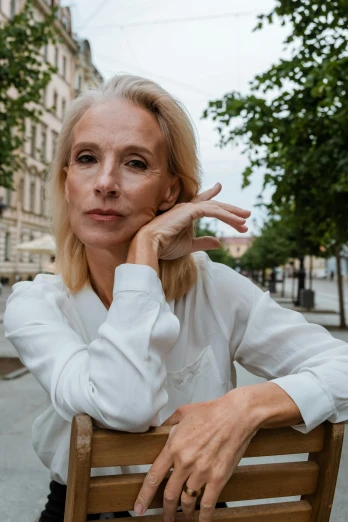 a woman sitting on top of a wooden chair, by Ulrika Pasch, pexels contest winner, photorealism, wearing white shirt, in a city square, hand on her chin, blonde swedish woman