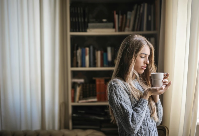 a woman standing in front of a window holding a cup, trending on pexels, reading in library, cosy, grey, coffee smell