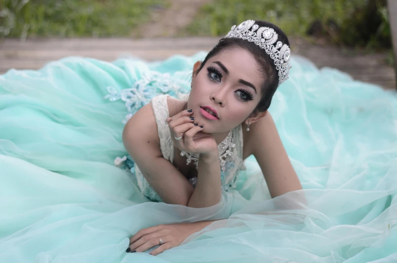 a woman in a blue dress laying on the ground, inspired by reyna rochin, pixabay, aestheticism, wearing tiara, south east asian with long, avatar image, 18 years old