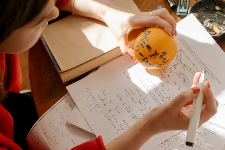 a woman sitting at a table writing on a piece of paper, by Joe Bowler, pexels contest winner, academic art, holding a tangerine, equations, holding a ball, formulae