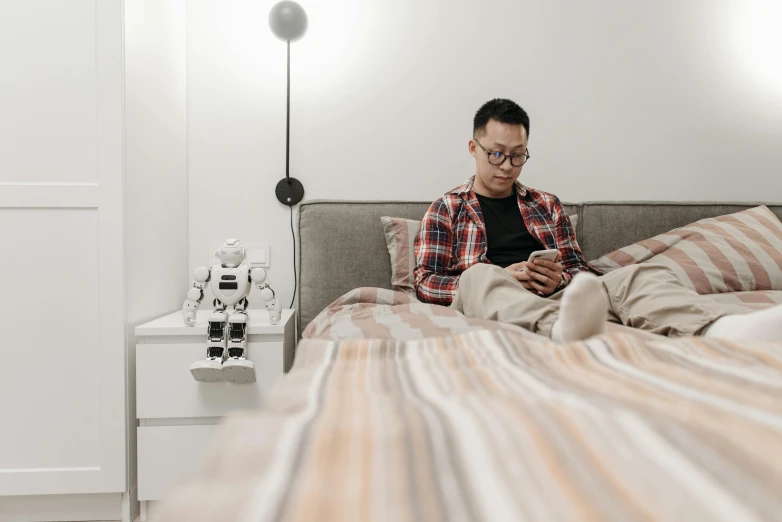 a man sitting on a bed using a cell phone, by Adam Marczyński, trending on pexels, pixel art, robot cyborgs, asian descent, family friendly, man with glasses