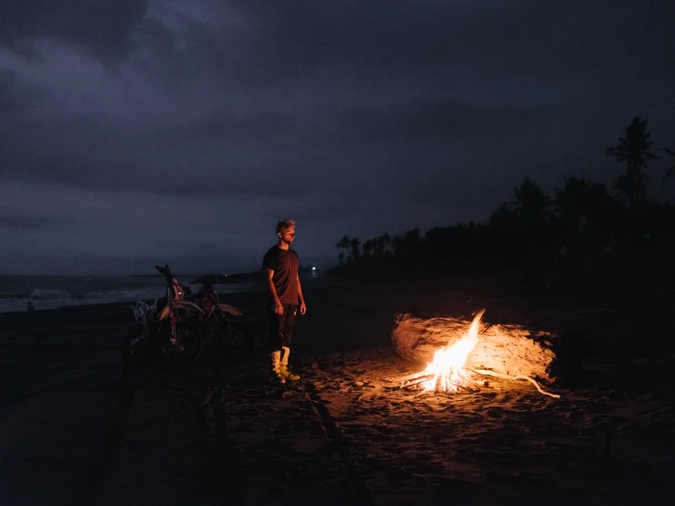 a man standing next to a fire on a beach, pexels contest winner, rain lit, alana fletcher, telling stories, low iso