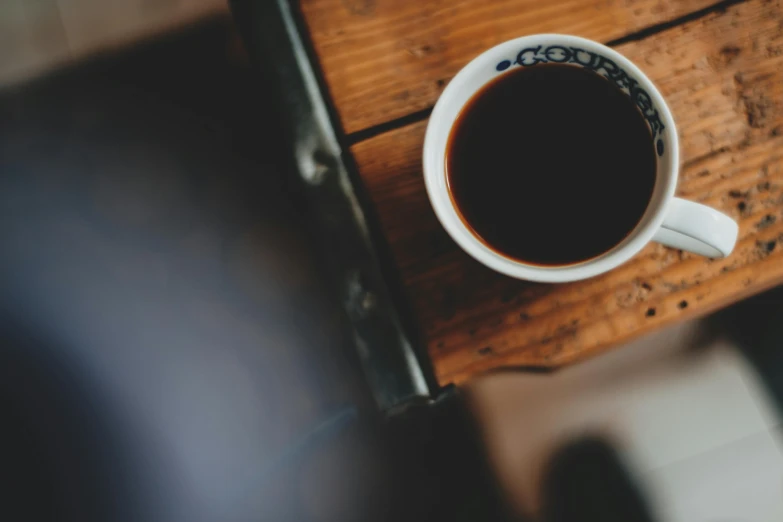 a cup of coffee sitting on top of a wooden table, by Niko Henrichon, trending on unsplash, fan favorite, background image, short focus depth, alessio albi